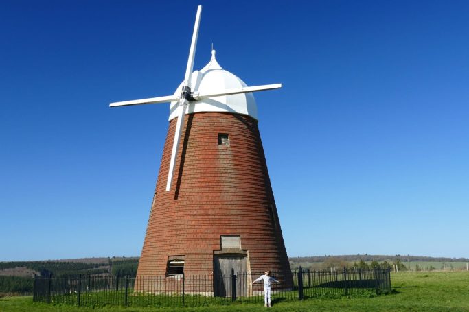 Red-brick windmill with white blades against a clear blue sky, surrounded by green grass.