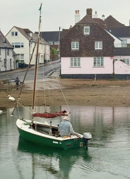 A small green boat is moored by a riverside with a man seated inside, near buildings.