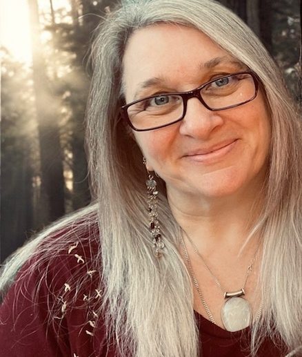 supervision Woman with long grey hair, glasses, and a warm smile in a forest setting.