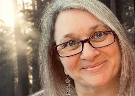 Smiling woman with glasses and long grey hair, surrounded by a forest backdrop.