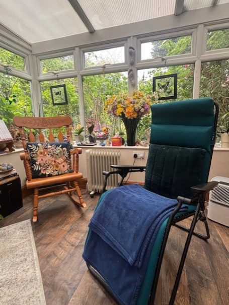 A cosy conservatory featuring a green chair, a rocking chair, and a vase of flowers.