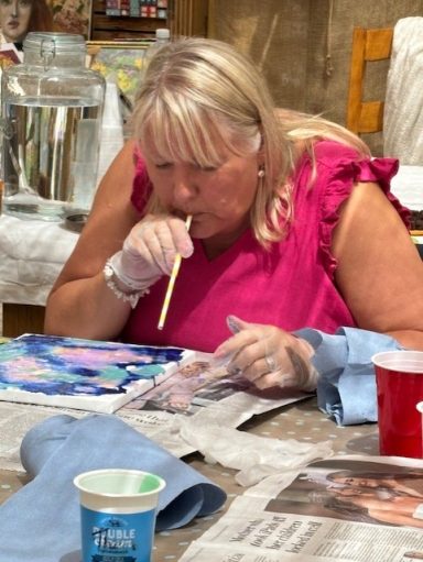 A woman in a pink top uses a paintbrush while sitting at a table covered in art supplies.