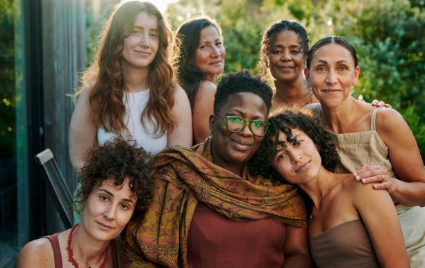 A diverse group of seven women posing together in a natural outdoor setting.