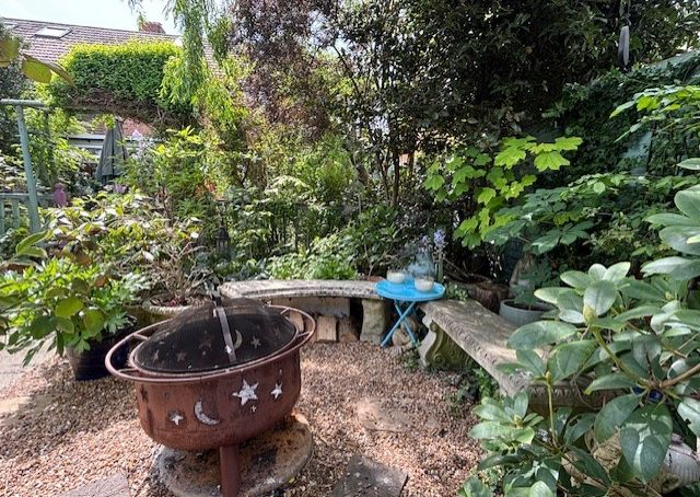 A vibrant garden scene with greenery, a rustic fire pit, and a wooden bench.