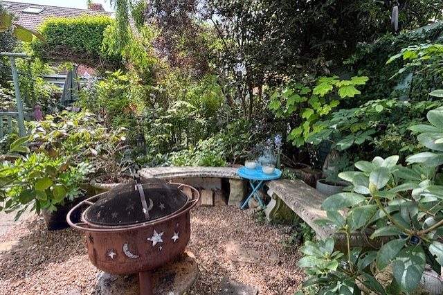 A rustic garden scene with a fire pit, surrounded by lush greenery and a wooden bench.