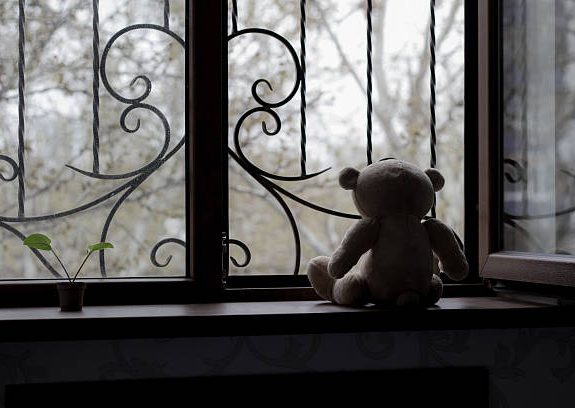 A teddy bear sits on a windowsill beside a small plant, with trees visible outside.