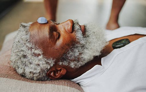 A man with grey hair lies down, a stone on his forehead, in a calming setting.