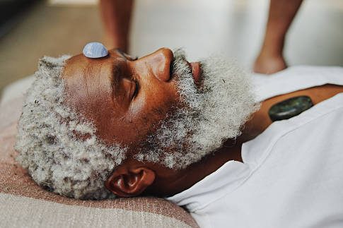 An older man relaxes with stones placed on his forehead and chest.
