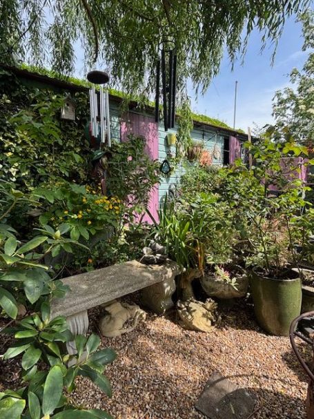 A vibrant garden with a stone bench, plants, and a wind chime against a colourful wall.