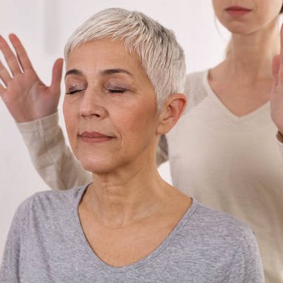 A calm, elderly woman with short grey hair, eyes closed, meditating with a person behind her.