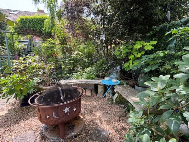 A rustic garden with a wooden bench, plants, and a decorative fire pit.