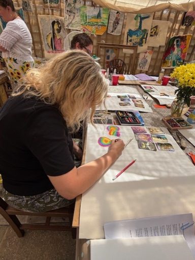 A person creates art at a table filled with colourful materials and flowers.