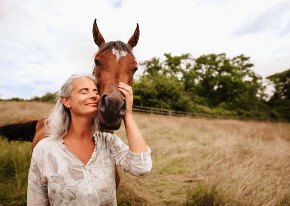 Smiling woman softly touching a horse's face in a grassy field.