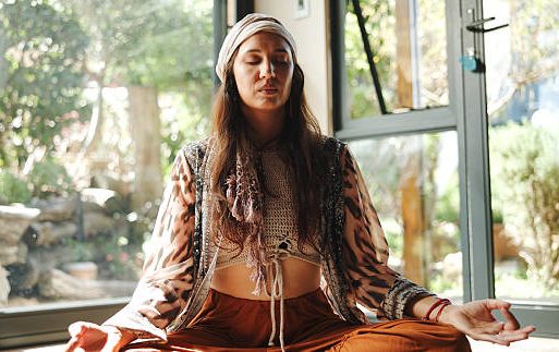 A woman meditating indoors, wearing headphones and a patterned top, with plants in the background.