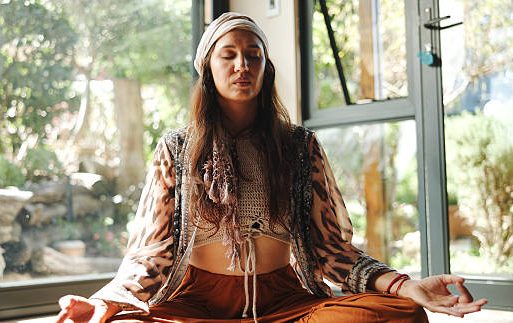 Woman meditating indoors, wearing a patterned top and headphones, with greenery in the background.