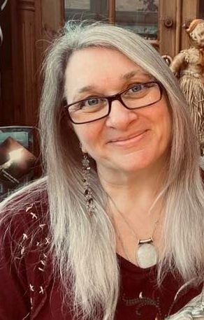 Smiling woman with long grey hair, wearing glasses and a pendant, against a wooden backdrop.