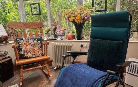 A sunlit room with a green chair, a rocking chair, and a vase of flowers on a table.